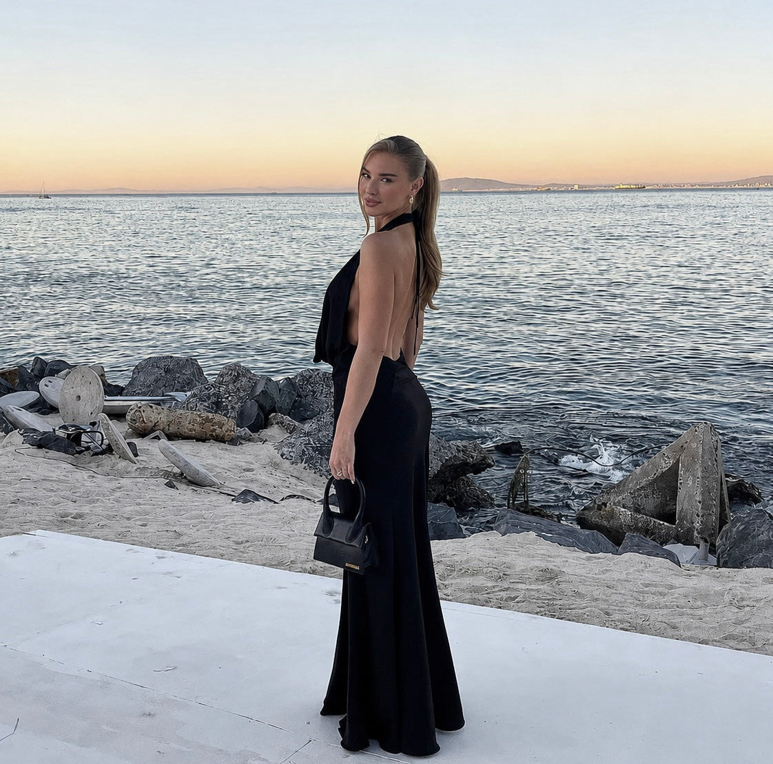 Woman in a black dress standing by the water at sunset